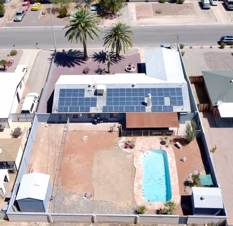 Aerial view of a single-story building with rooftop solar panels and an enclosed backyard containing a swimming pool, patio area, and palm trees by the street.