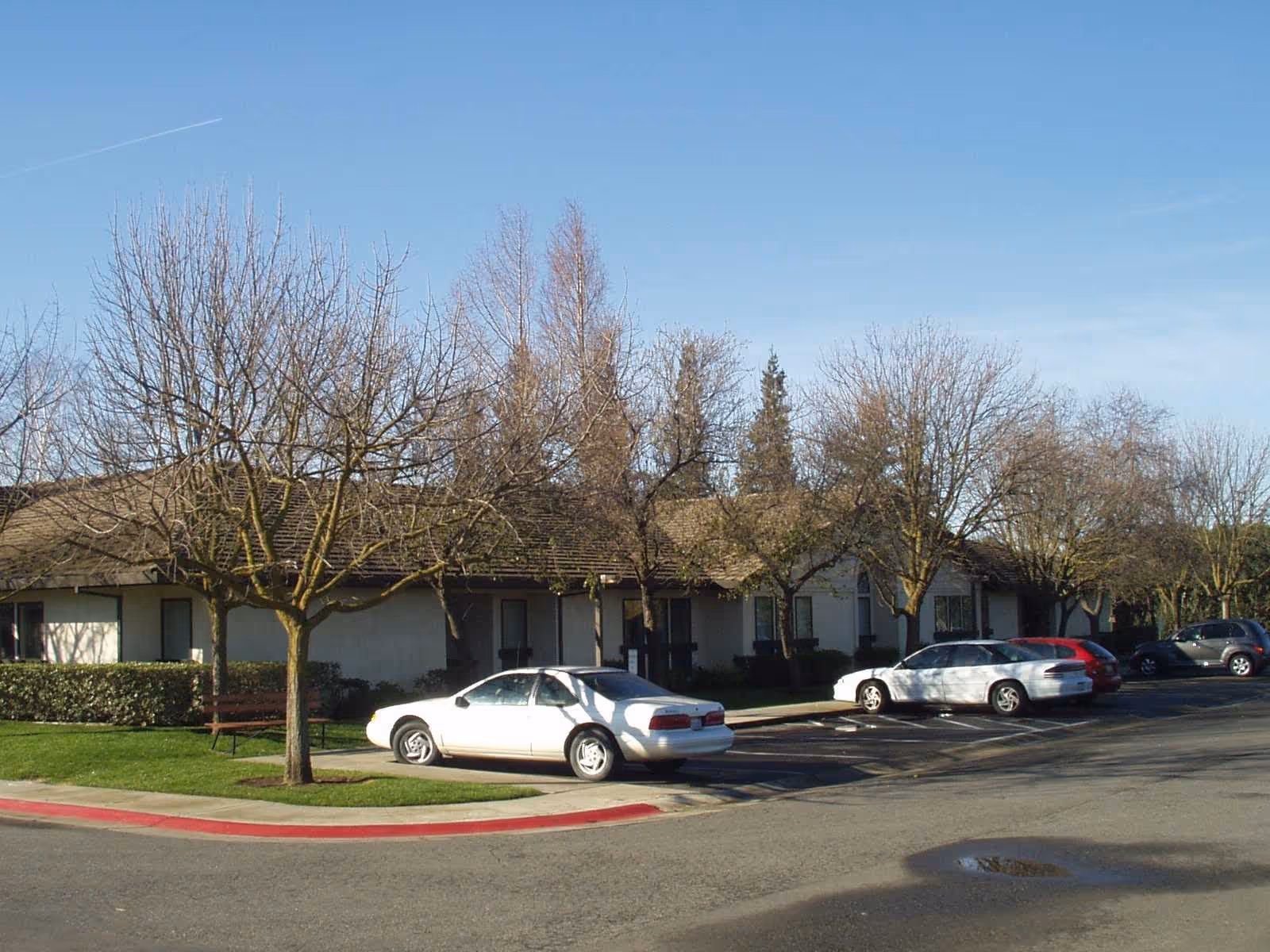 Single-story retirement village building front with leafless trees and several parked cars under a clear blue sky.