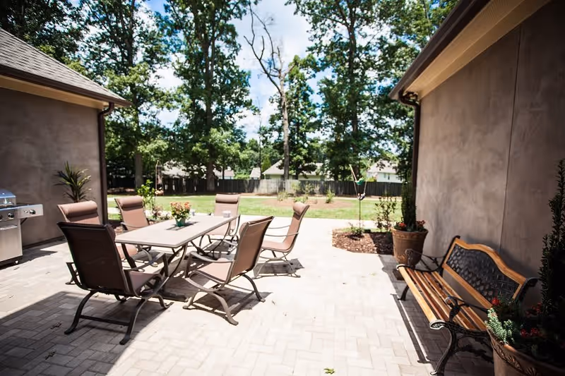 Outdoor patio area with a rectangular table surrounded by six chairs, a wooden bench with metal armrests, potted plants, a barbecue grill, and trees in the background under a partly cloudy sky.