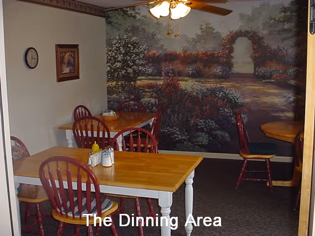 A dining area with wooden tables and red wooden chairs. The room features a large mural on the back wall depicting a garden scene with flowers and an archway. There is a ceiling fan with lights above the tables, and a clock and framed picture on the left wall.