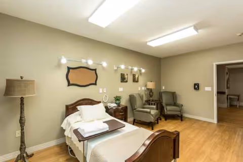 A furnished senior living bedroom with a bed, chairs, and warm lighting.