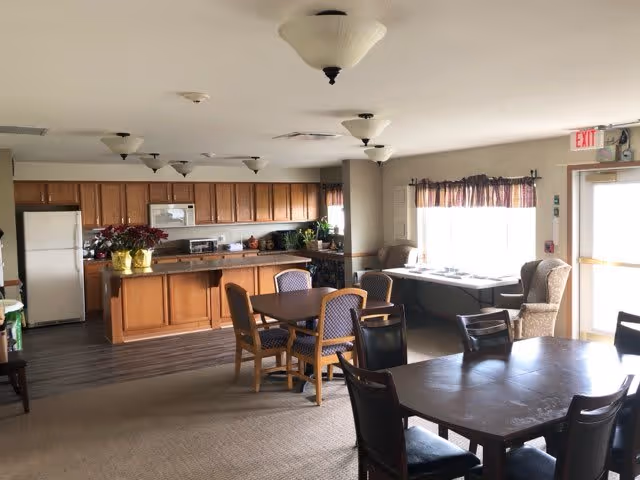 Communal dining area with several tables and chairs in front of a kitchenette with wooden cabinets and appliances.