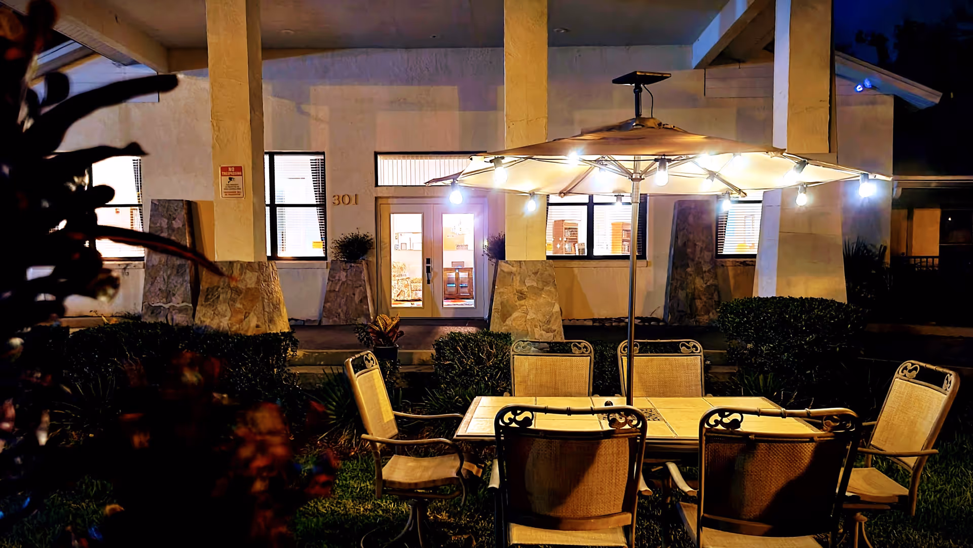 Outdoor patio area at night with a table and six chairs under a large umbrella adorned with string lights. The background shows the entrance to a building with windows and a door illuminated from inside.