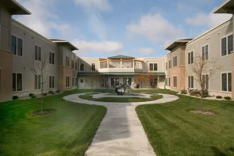 Outdoor courtyard area of Oak Park Place Madison with a circular walkway, green grass, young trees, and a central sculpture, surrounded by a two-story building under a partly cloudy sky.