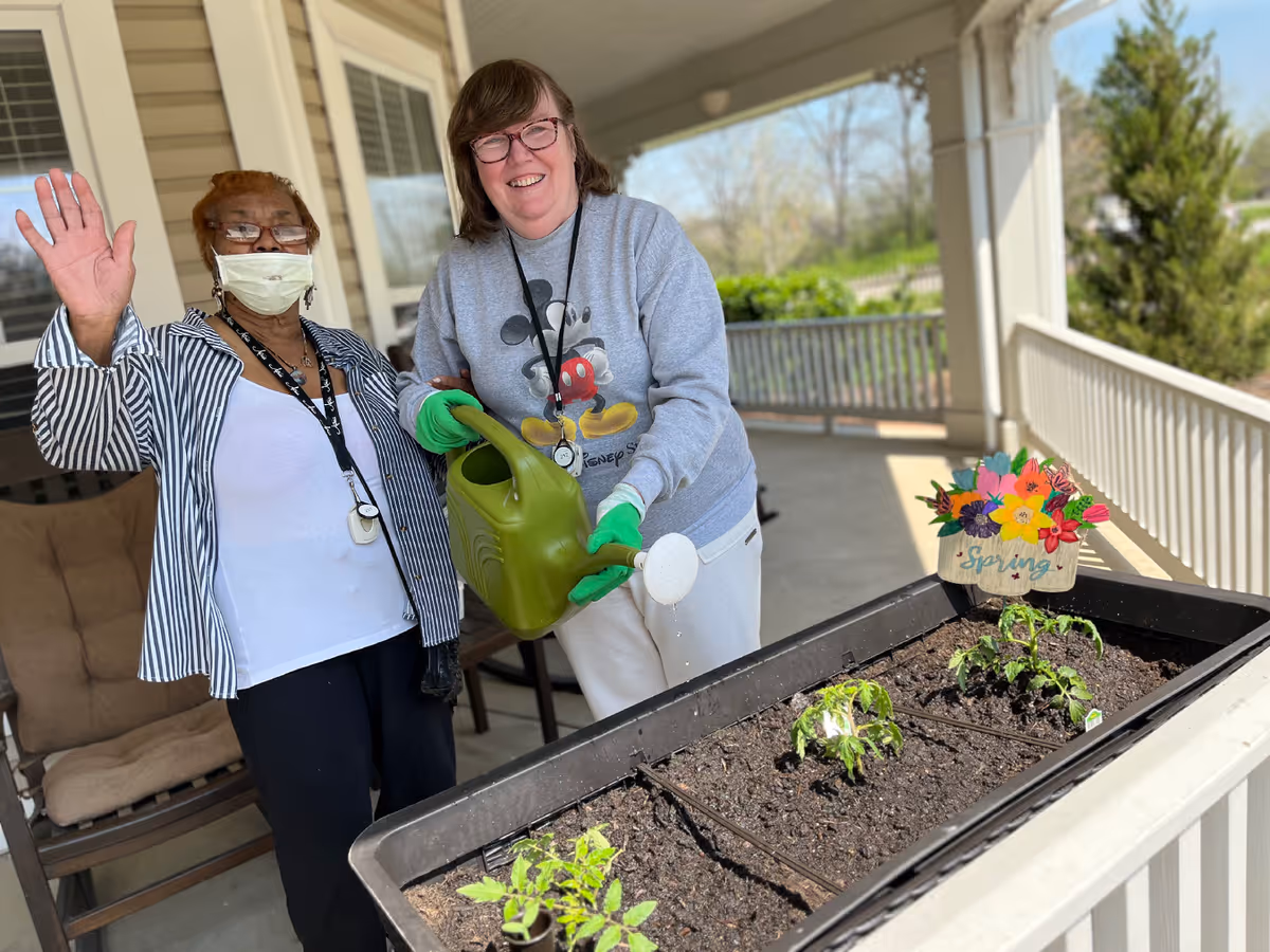 Two women on a covered porch tending a planter box, one waving and the other pouring water from a green watering can.
