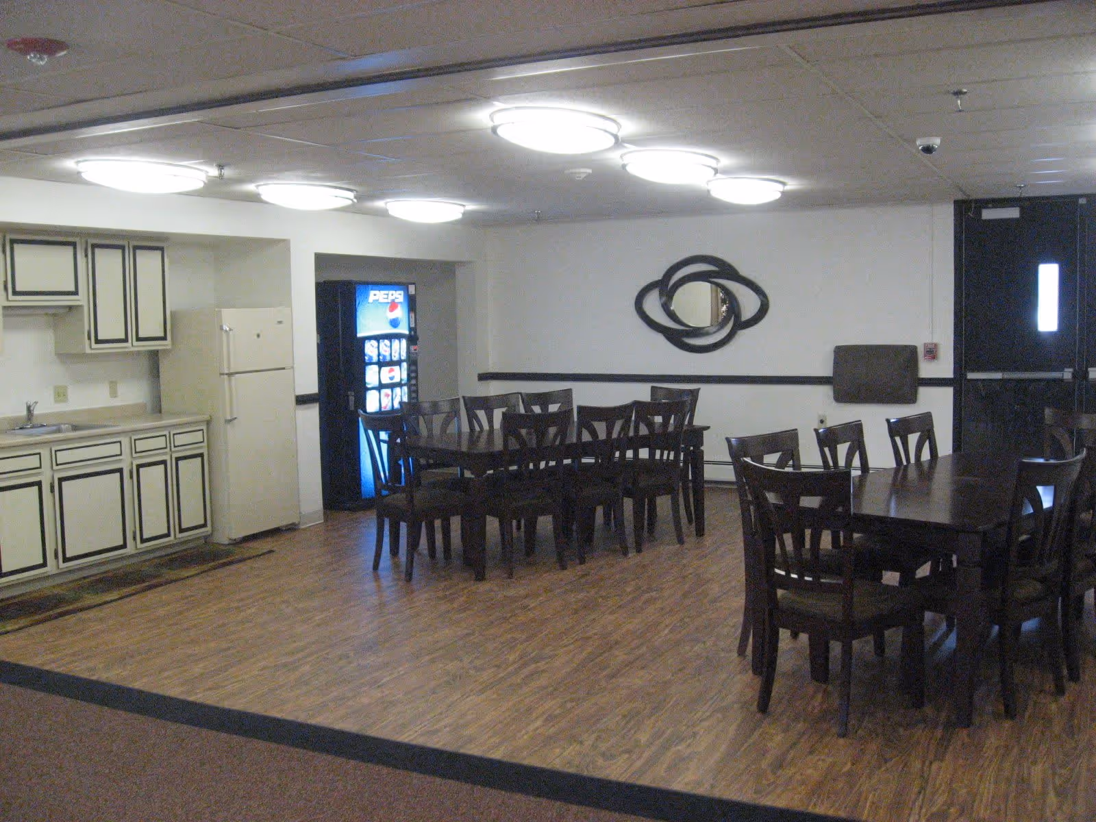 Interior view of a communal dining area with wooden tables and chairs, a kitchenette with cabinets and a refrigerator, and a Pepsi vending machine in the corner. The room has wood flooring, white walls, and ceiling lights.
