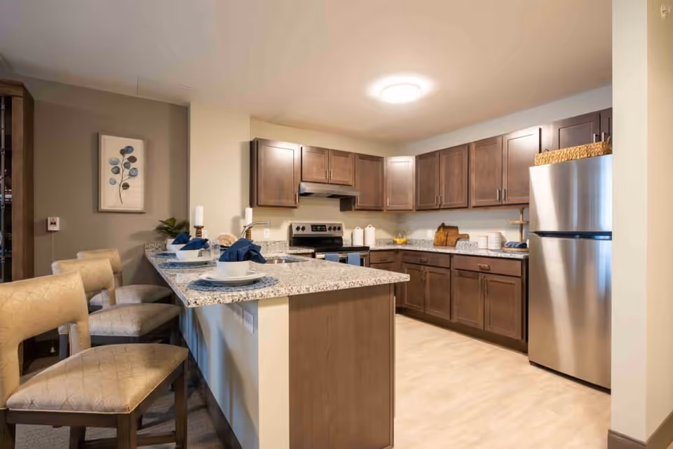 A modern kitchen with dark wood cabinets, a stainless steel refrigerator, an electric stove, and a granite countertop island with three beige cushioned chairs. The countertop is set with white plates and blue napkins. The kitchen has light-colored flooring and a ceiling light fixture.