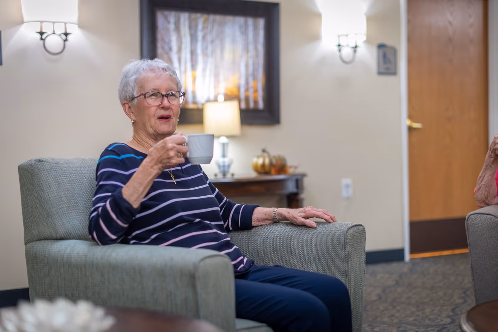 An elderly woman with short white hair and glasses sits comfortably in a green armchair, holding a white mug. She is wearing a navy blue and white striped long-sleeve shirt and dark pants. Behind her, there is a wooden table with a lamp and decorative items, and a framed picture on the wall. The room has soft lighting and a closed wooden door is visible in the background.