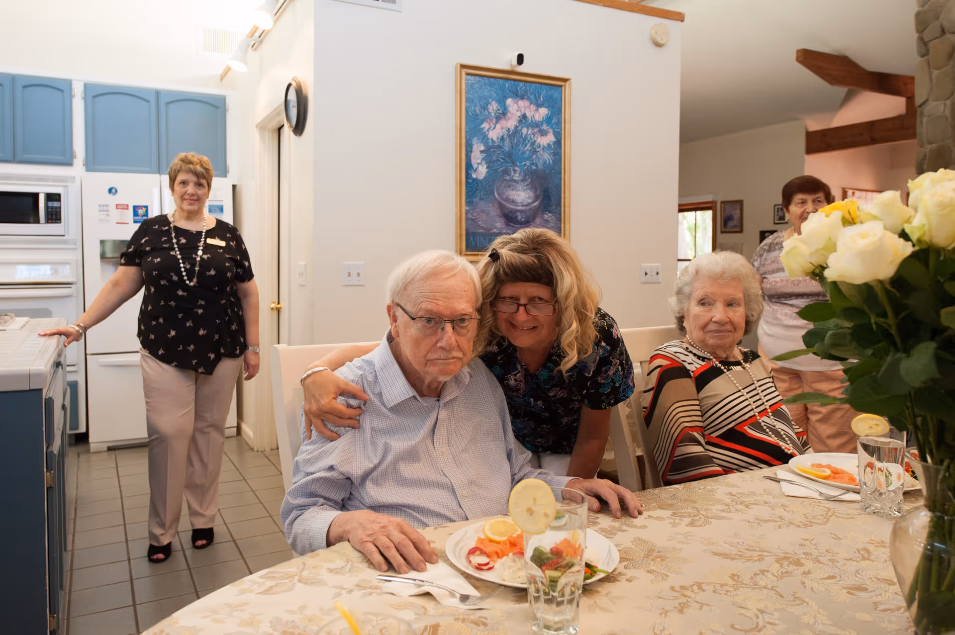 A group of elderly people and a caregiver gathered around a dining table in a kitchen area. One elderly man is seated at the table with a plate of food and a glass of water with a lemon slice, while a woman leans in to hug him. Another elderly woman is seated at the table, and two other women stand in the background near the kitchen appliances. The room has light-colored walls, blue kitchen cabinets, and a floral painting on the wall.