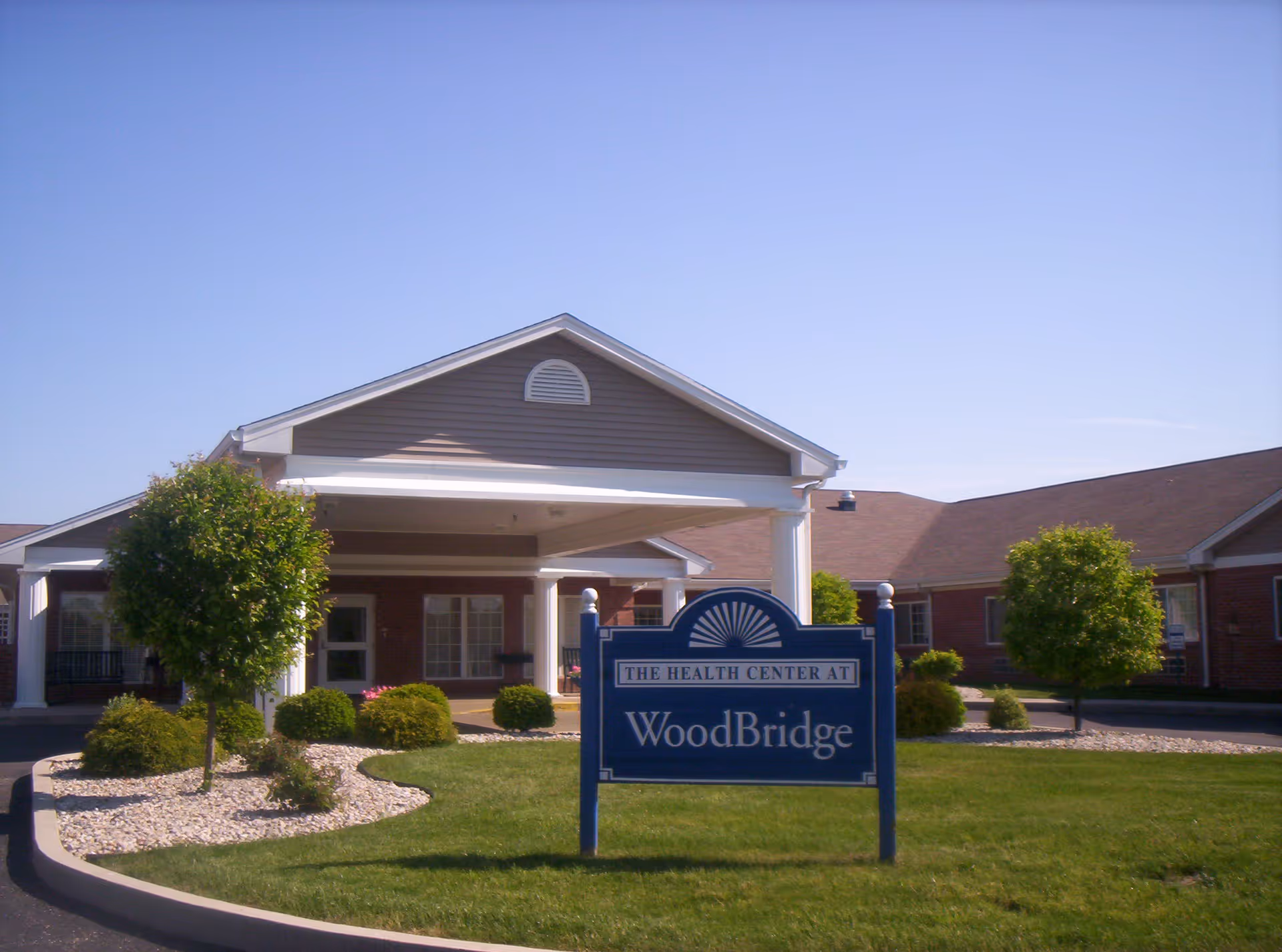 Front entrance of WoodBridge Health Campus showing a covered drop-off with columns, landscaping, and a blue sign reading "WoodBridge".