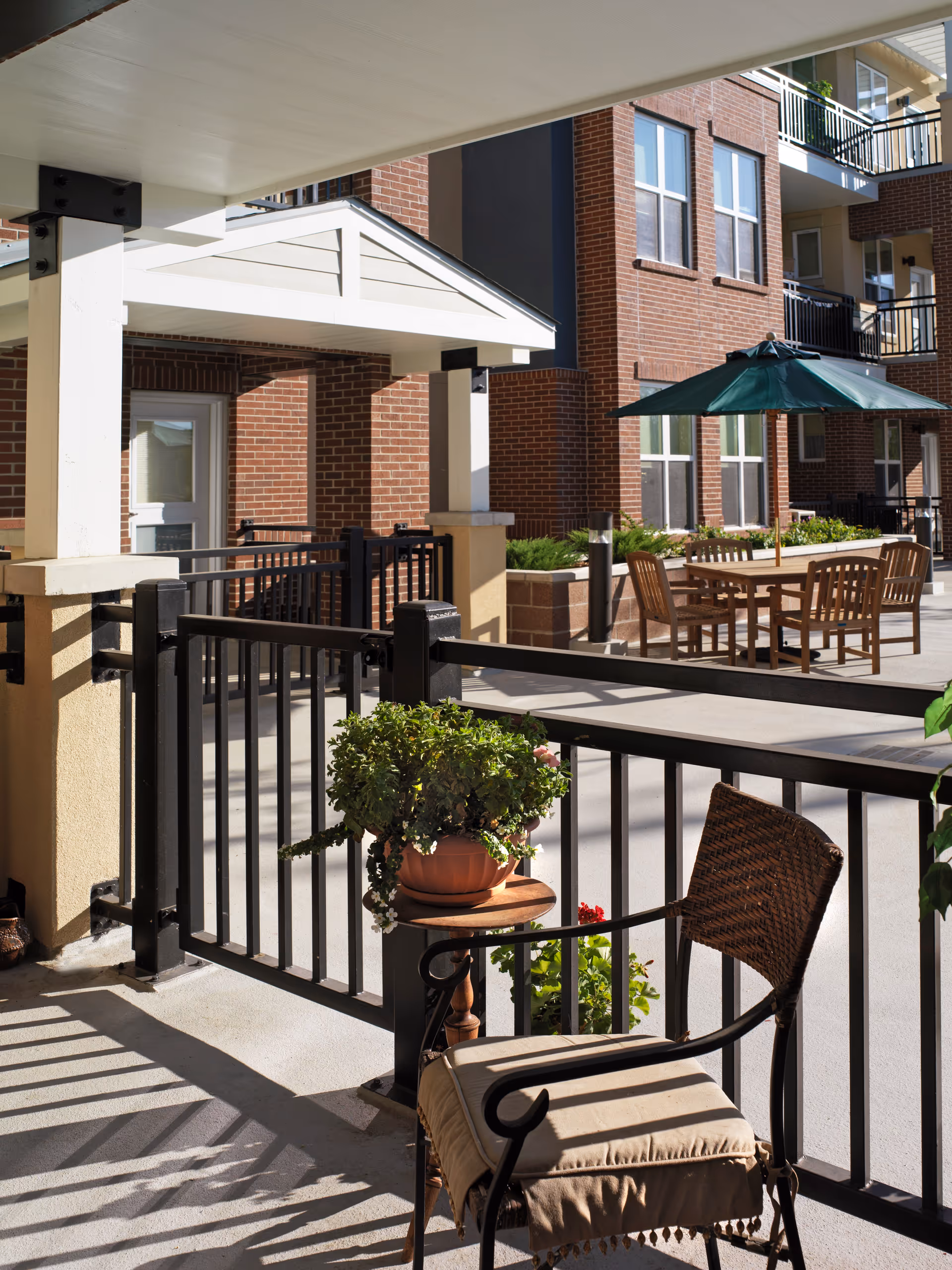 Outdoor patio area at a senior living facility with a cushioned wicker chair and a small round table holding a potted plant in the foreground. In the background, there is a wooden table with chairs and a green umbrella, surrounded by brick buildings with multiple windows and balconies.