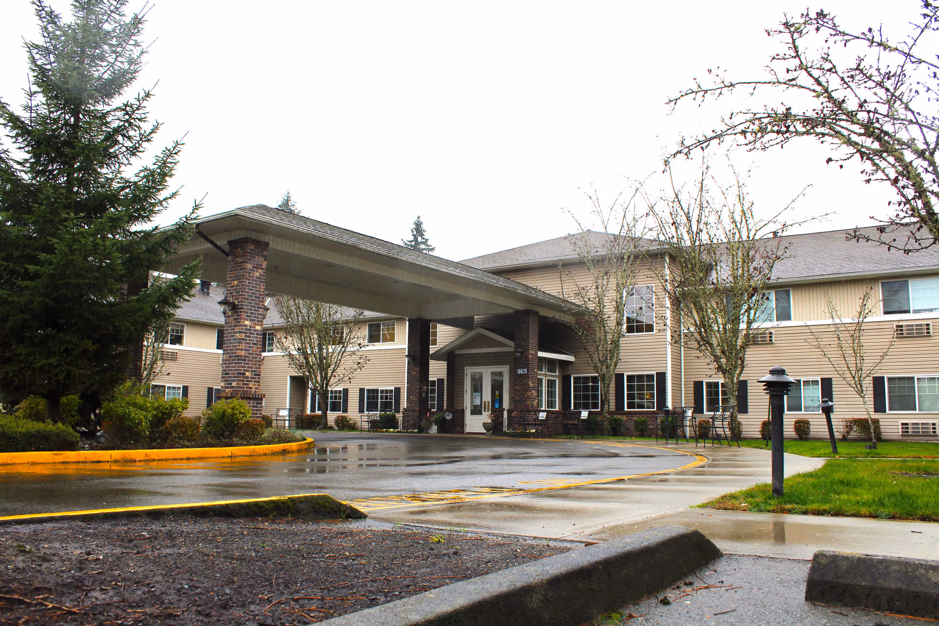 Exterior view of Normandy Park Senior Living facility showing a two-story building with beige siding and brick pillars supporting a covered entrance. The driveway and sidewalk are wet from rain, and there are trees and shrubs around the entrance area.