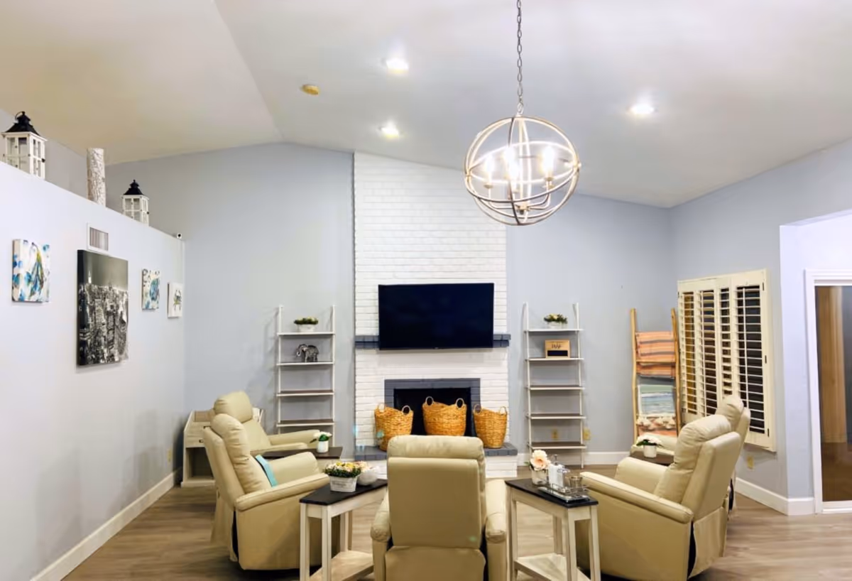 Bright communal living room with beige recliner chairs arranged around a TV mounted above a white brick fireplace.