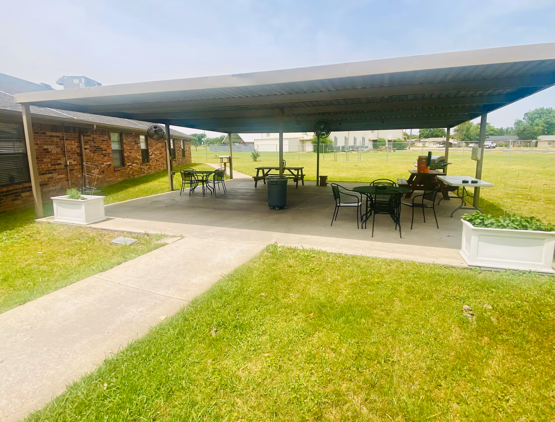 Outdoor covered patio area with several tables and chairs, a picnic table, and a barbecue grill. The patio is adjacent to a brick building and surrounded by a grassy lawn and fenced yard.