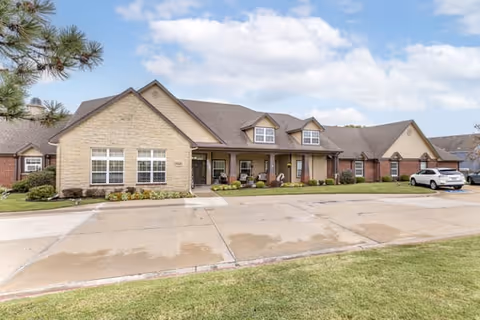 Exterior view of a single-story senior living facility building with a combination of stone and brick facade, multiple windows, and a covered porch area with seating. There is a driveway and parking area in front, along with a grassy lawn and a tree on the left side. The sky is partly cloudy.