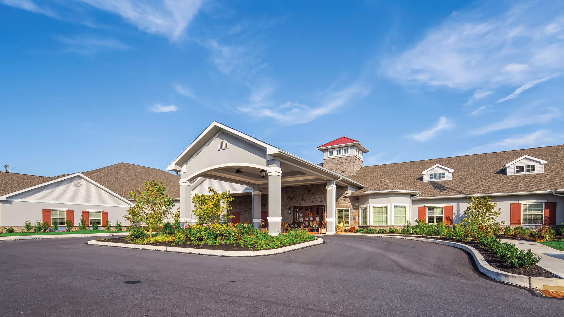 Front exterior view of the Legend of Lancaster facility showing a large driveway with a covered entrance, landscaped greenery, and a building with beige walls, red shutters, and a stone tower under a blue sky with some clouds.