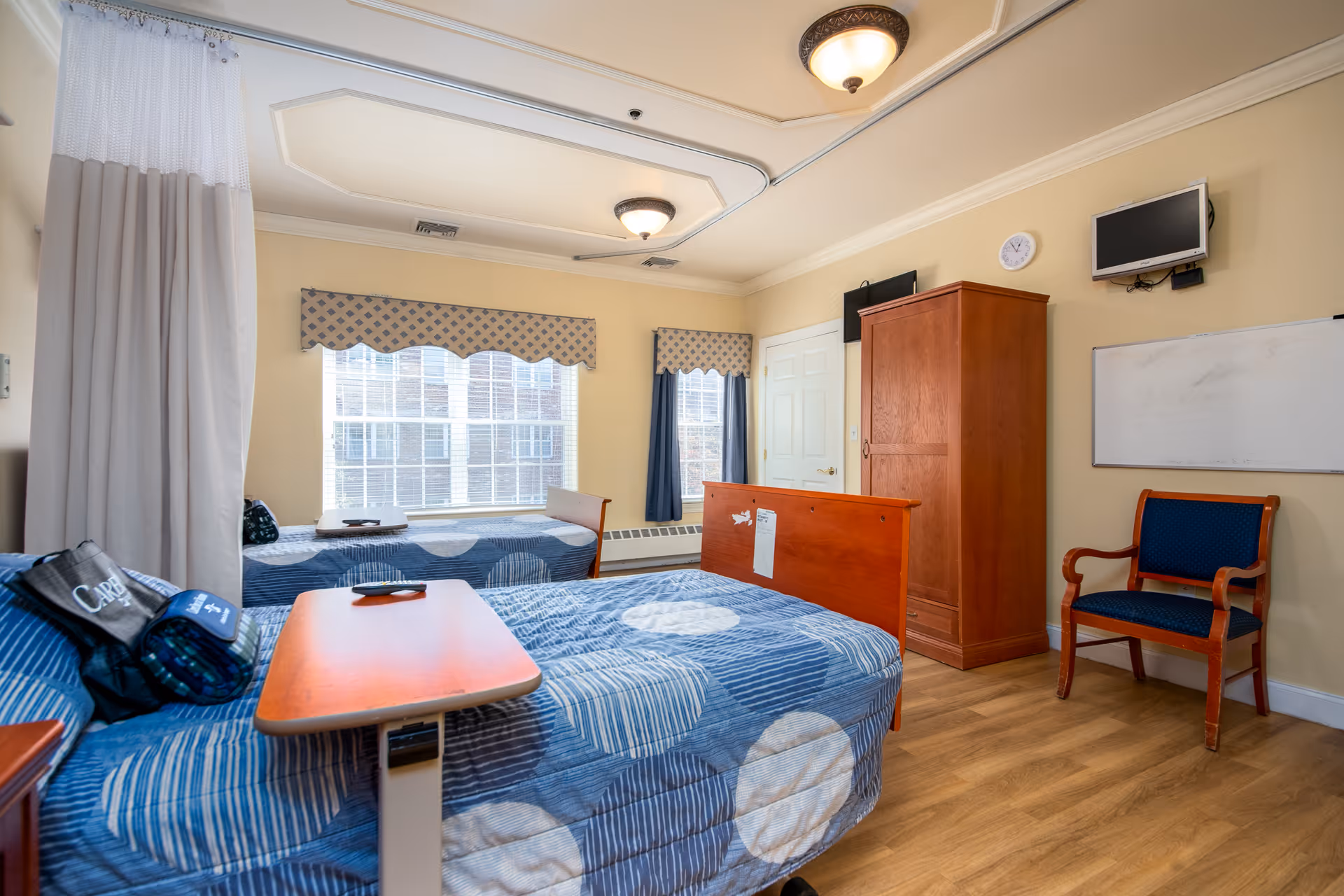 A bright and clean bedroom in a senior living facility with two beds covered in blue patterned bedspreads. The room features a wooden wardrobe, a blue upholstered chair, a wall-mounted TV, a whiteboard, and large windows with patterned valances letting in natural light. There is a ceiling light fixture and a privacy curtain partially visible on the left side.