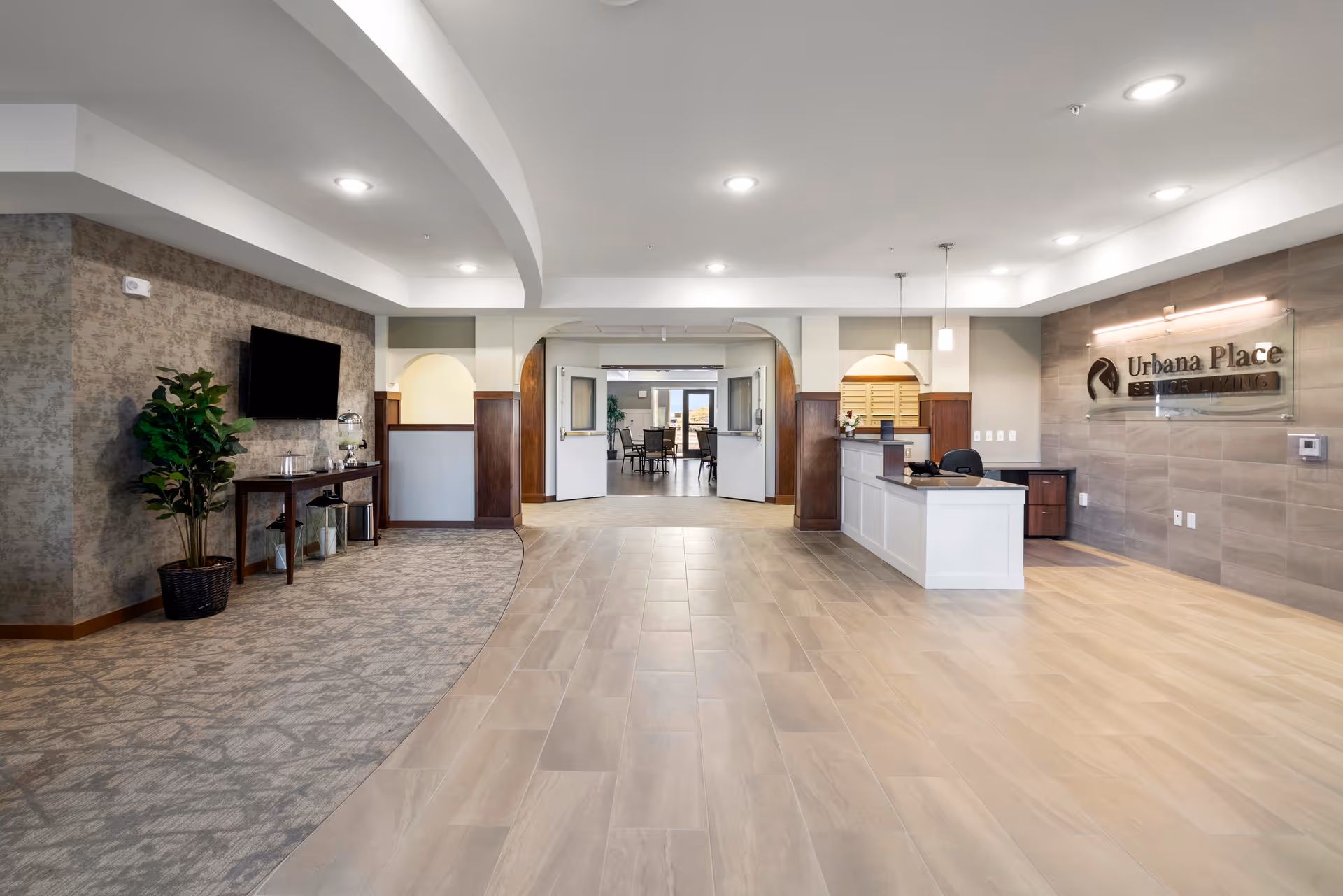Reception area of Urbana Place Senior Living featuring a front desk on the right with a sign on the wall, a seating area with a TV and plant on the left, and an open doorway leading to a dining area in the background.