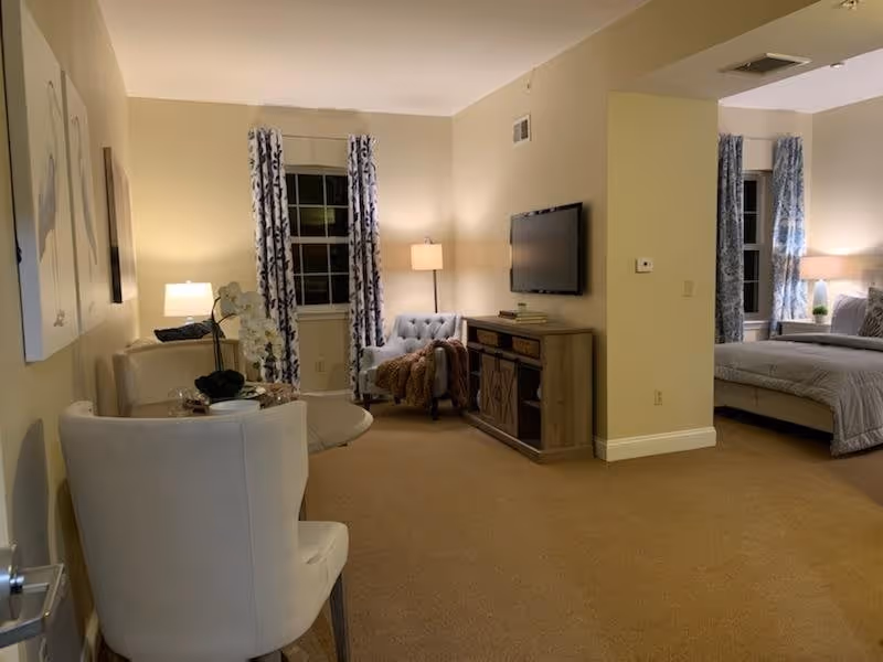 Interior view of a senior living facility room with a small dining table and two chairs on the left, a cozy sitting area with an armchair and floor lamp in the center, and a bedroom area with a bed and nightstand visible on the right. The walls are painted light beige, and windows have patterned curtains.
