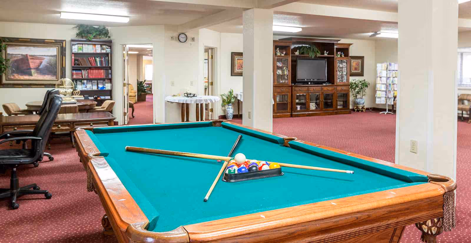 An indoor common room with a pool table in the foreground, seating, a TV cabinet and bookshelves in the background.