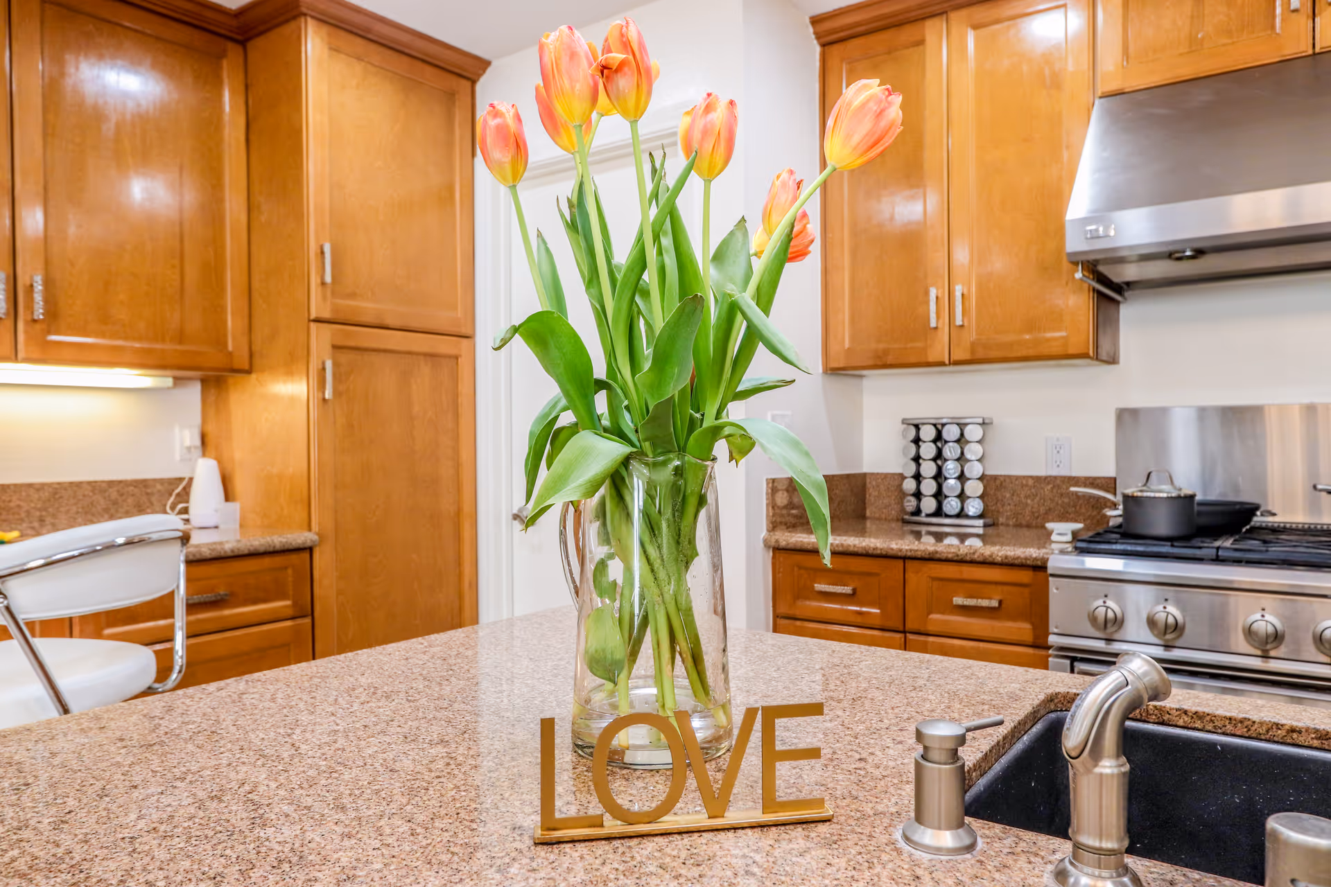 A modern kitchen with wooden cabinets, a granite countertop island with a vase of orange tulips and a decorative 'LOVE' sign, a stainless steel stove with pots, and a sink with a faucet and soap dispenser.