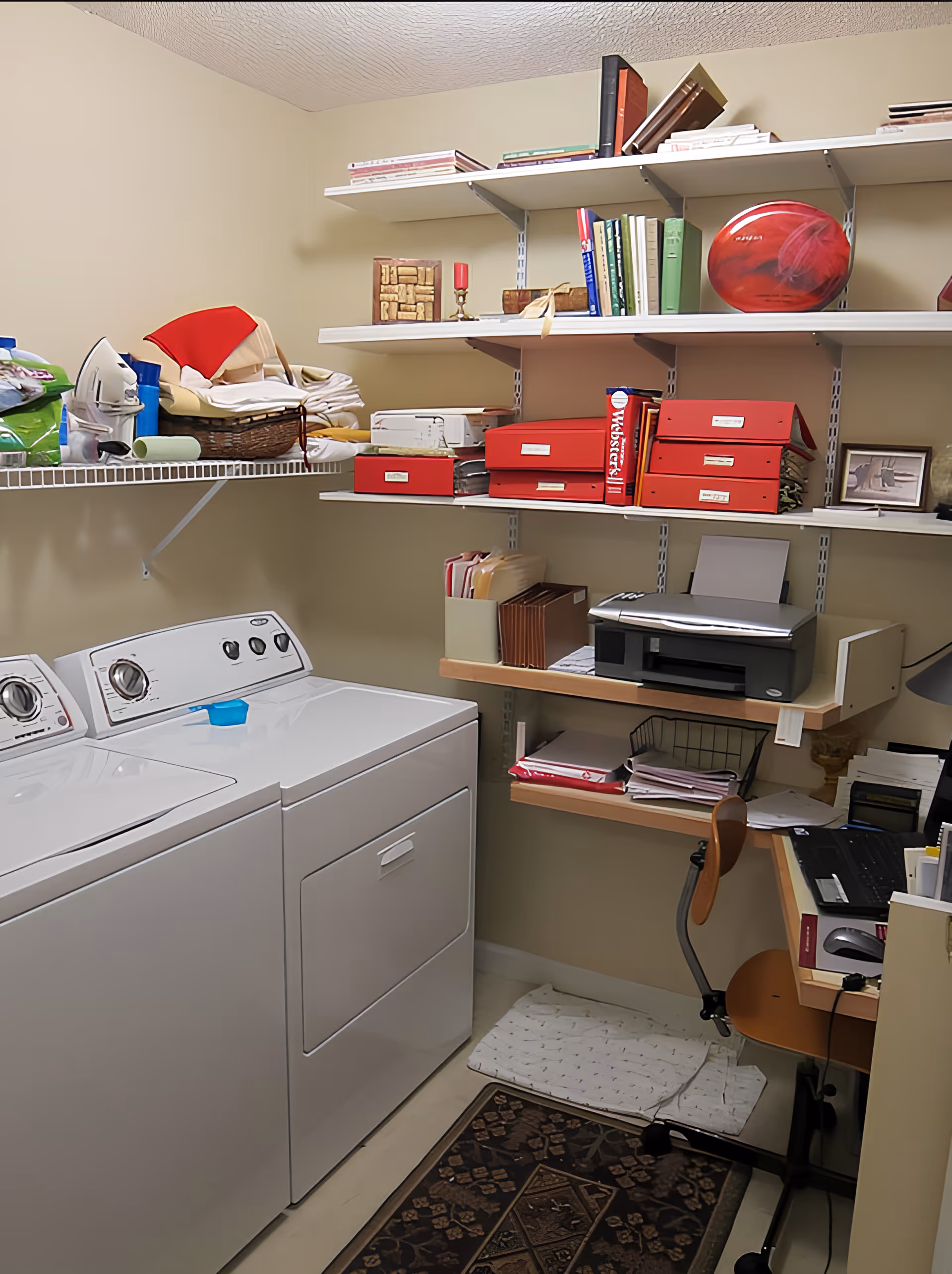 A laundry room with a white washing machine and dryer side by side. Above them are shelves holding folded linens, an iron, and various household items. On the right side, there is a small office area with a desk, chair, printer, and organized files and books on shelves.