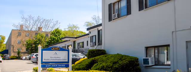 Exterior view of West Roxbury Health & Rehabilitation Center building with a parking lot, greenery, and a sign displaying the facility name and contact number.
