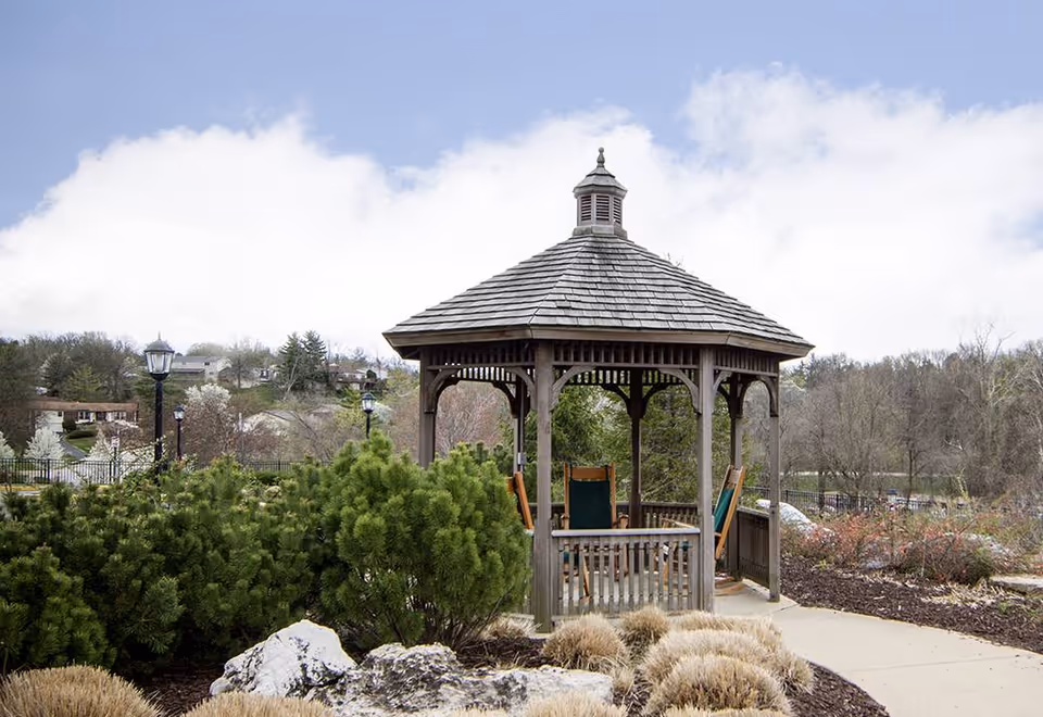 A wooden gazebo with a shingled roof and green cushioned chairs inside, situated in a landscaped garden area with shrubs, rocks, and a paved walkway. In the background, there are trees and houses under a partly cloudy sky.