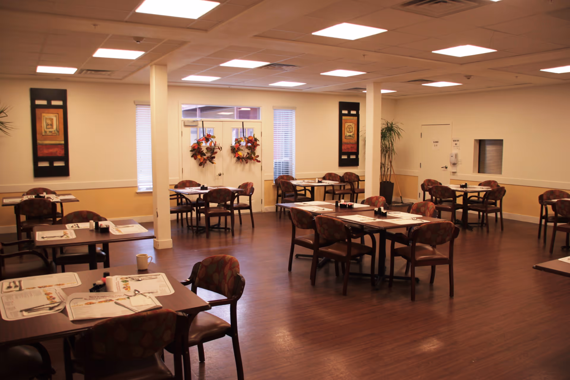 Spacious assisted living dining room with multiple set tables and chairs under ceiling lights.
