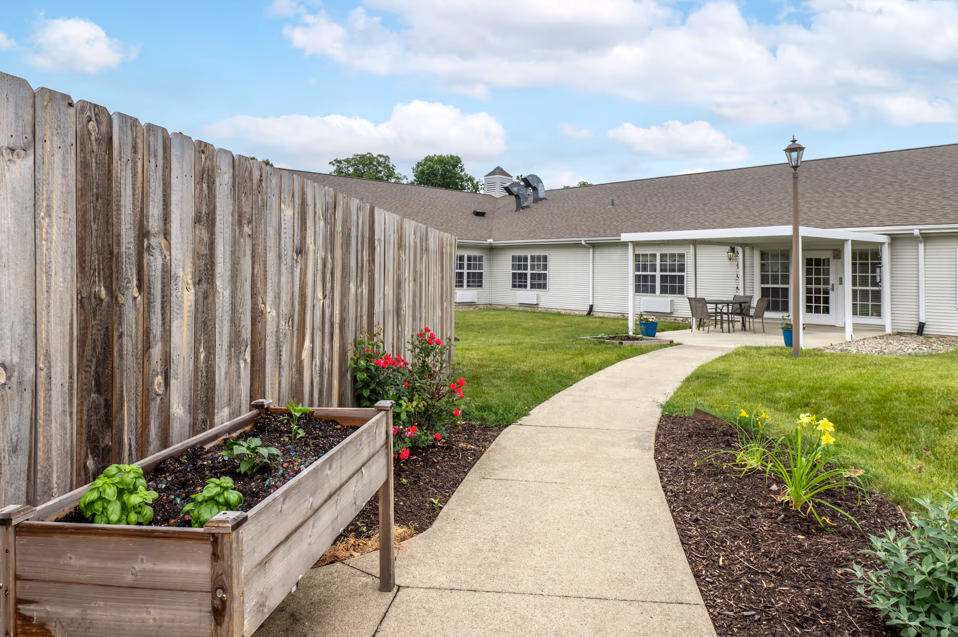Outdoor garden area at The Terrace at Ft Wayne featuring a raised wooden planter box with green plants, a wooden fence, a concrete walkway leading to a building with white siding and multiple windows, a patio table with chairs, and a lamppost. The sky is partly cloudy.