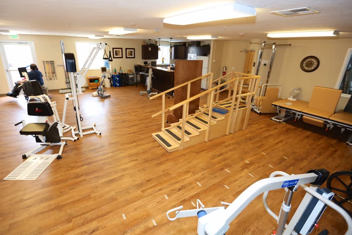 A spacious rehabilitation room with wooden flooring featuring exercise equipment, a set of wooden stairs with handrails for physical therapy, padded therapy tables, and a small kitchen area in the background. A person is seated on an exercise machine near the door.