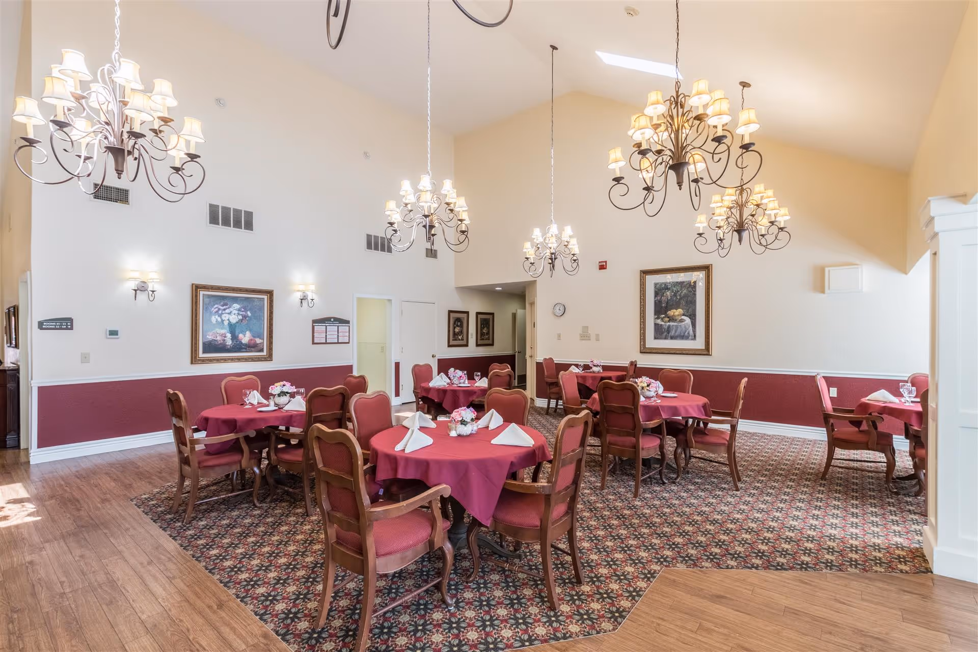 A dining room with several round tables covered with red tablecloths and set with white napkins and glassware. The room features patterned carpet, wooden chairs with red upholstery, multiple ornate chandeliers hanging from a high ceiling, framed artwork on the walls, and a combination of wood and carpet flooring.