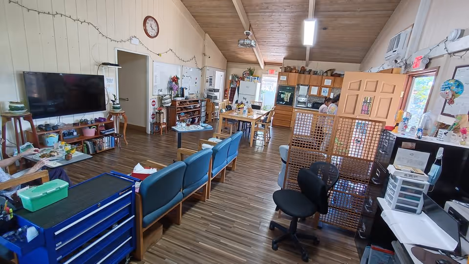 Interior view of a senior living facility common area with wooden floors and a vaulted wooden ceiling. The room includes a row of blue chairs, a large flat-screen TV mounted on the wall, bookshelves, a dining table with chairs, and a kitchen area in the background. There are various decorations, plants, and office supplies visible, along with a clock on the wall and a room divider near the kitchen.