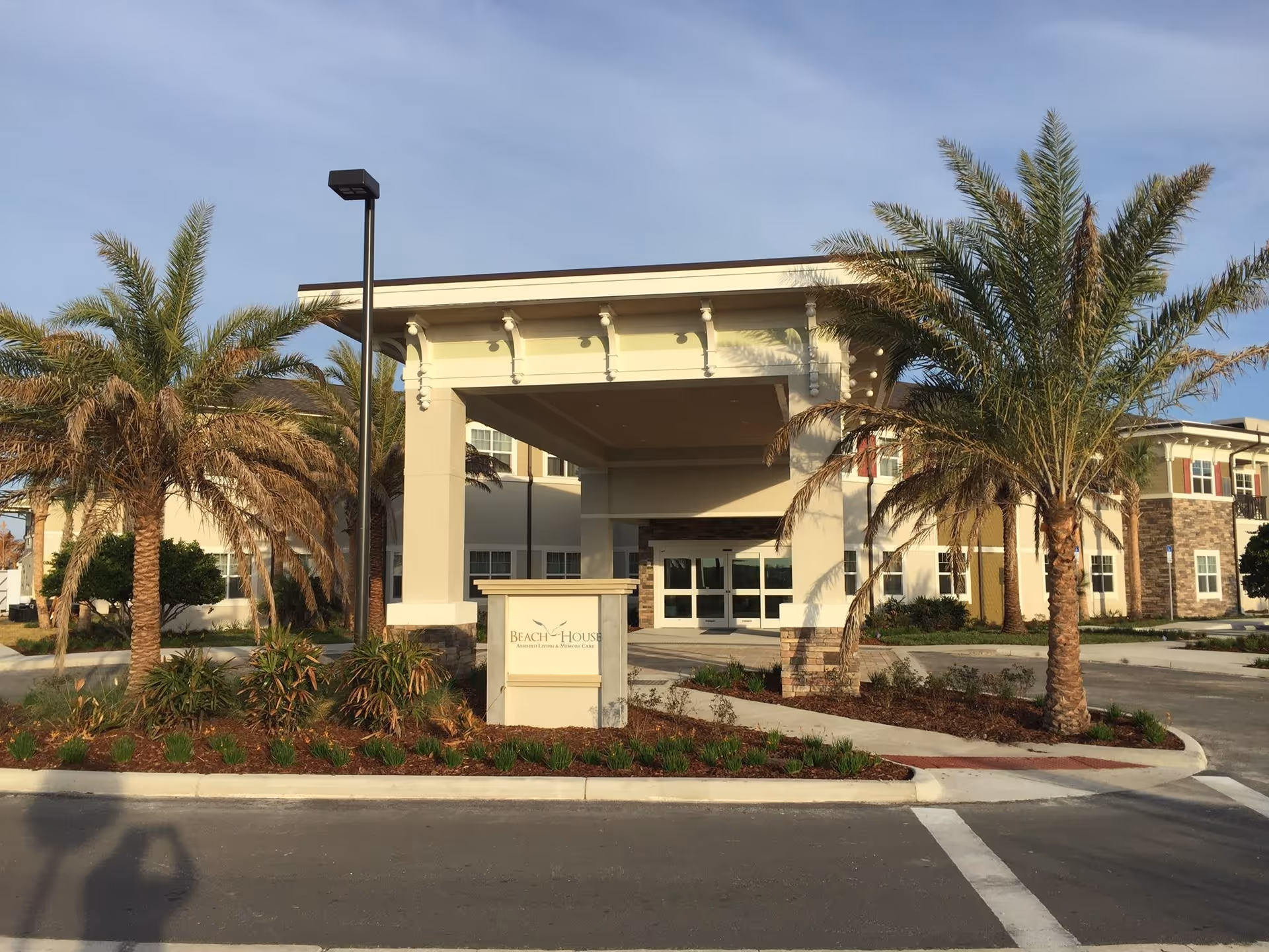 Front exterior view of Beach House Assisted Living & Memory Care facility with a covered entrance, palm trees, landscaped garden, and clear sky.