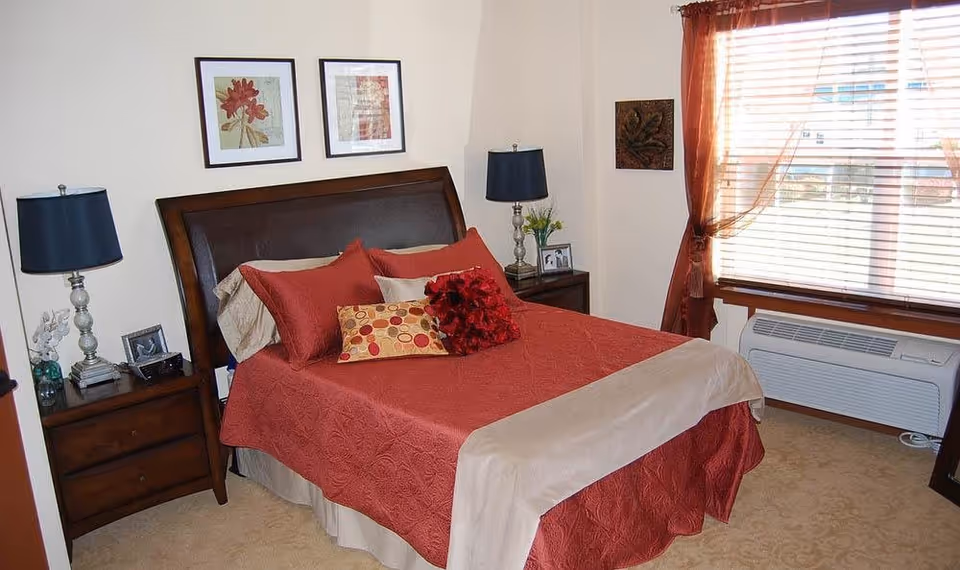 A cozy bedroom with a double bed covered in a red quilt and decorative pillows. The bed has a dark wooden headboard and is flanked by two matching nightstands, each with a lamp. There are two framed floral pictures hanging above the bed. A window with wooden blinds and sheer red curtains is on the right side of the room, with an air conditioning unit below it.