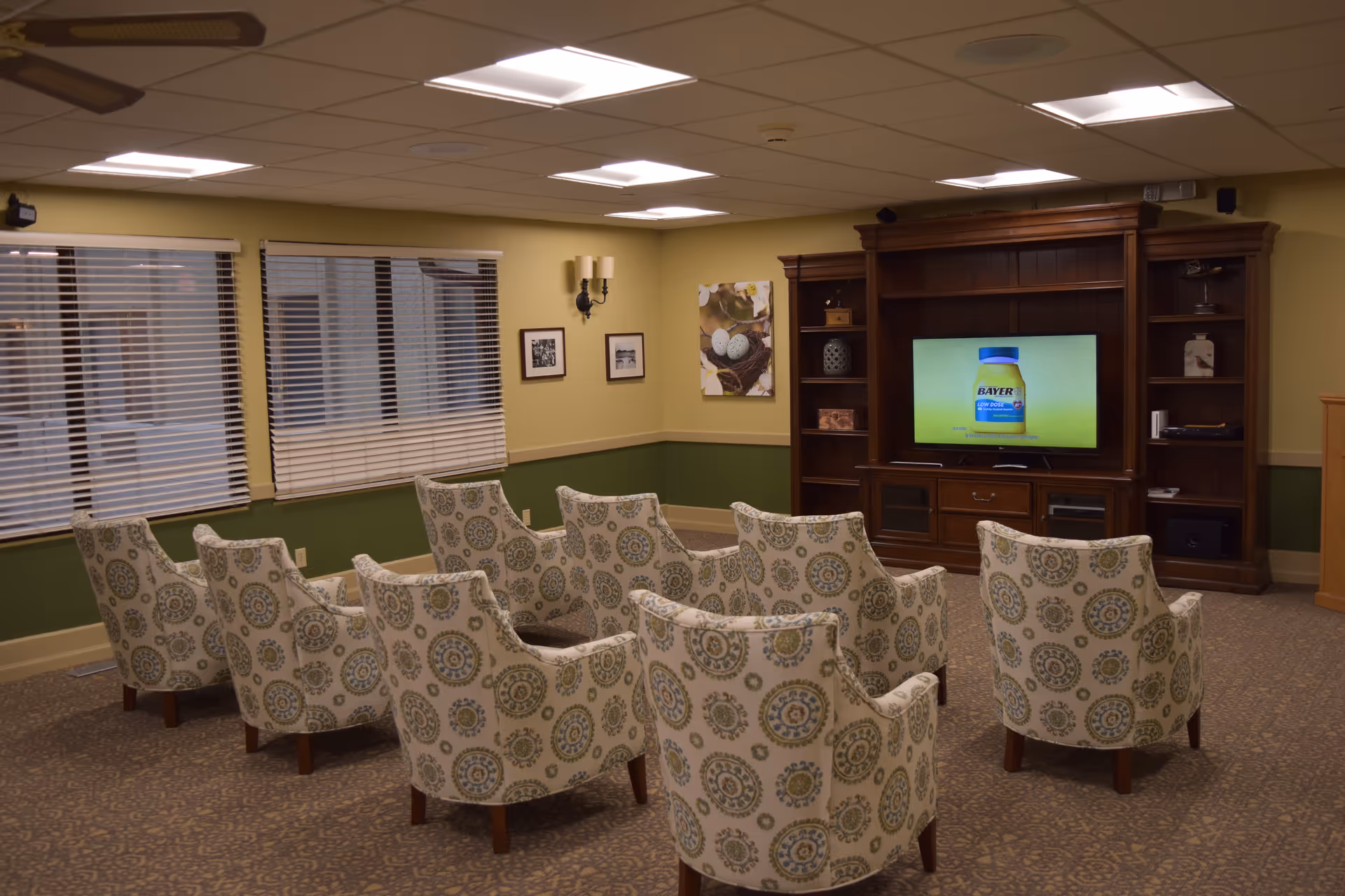 A cozy common area with patterned armchairs arranged in rows facing a large wooden entertainment center with a flat-screen TV. The room has green and beige walls, carpeted floor, windows with blinds, and ceiling lights.