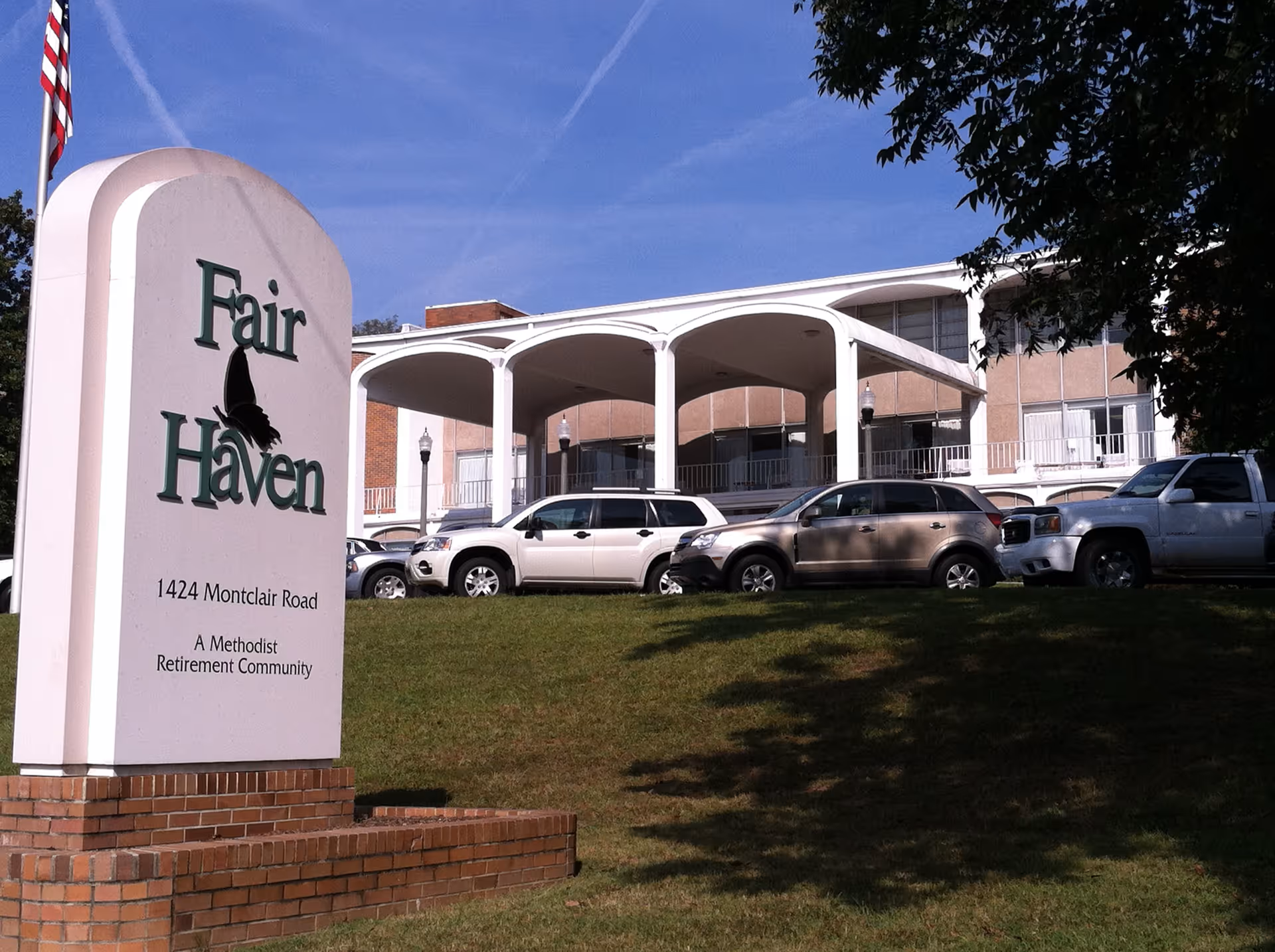 Exterior view of Fair Haven, a Methodist retirement community, showing the main building with a covered entrance and several parked cars on a grassy hill. A large sign with the community's name and address is visible in the foreground.