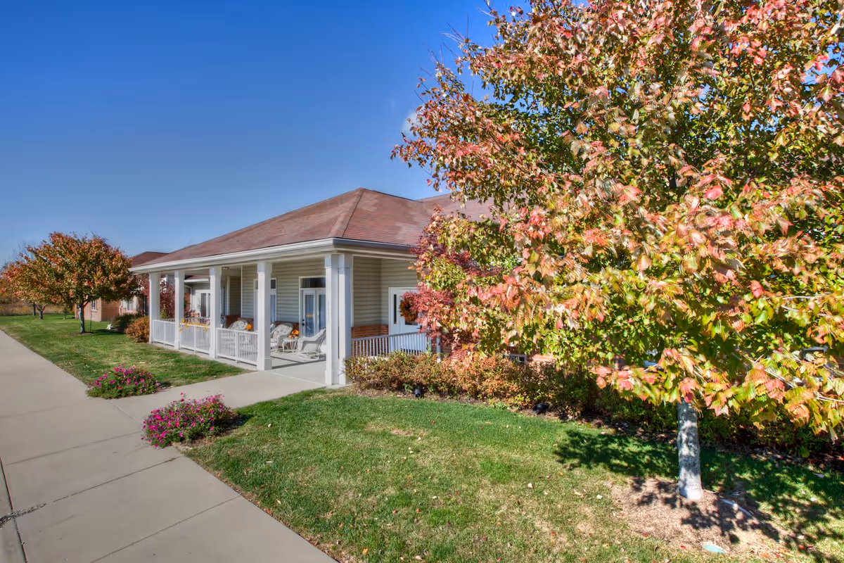 Exterior view of a single-story building with a covered porch featuring white railings and chairs. The building is surrounded by green grass, flowering plants, and trees with autumn-colored leaves under a clear blue sky.