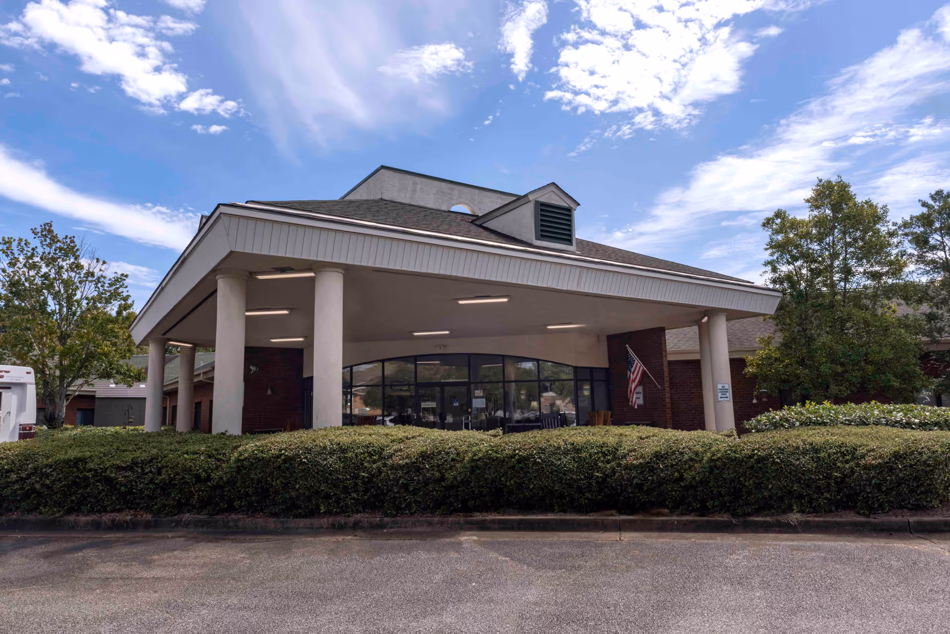 Front entrance of a brick assisted living facility with a covered porte-cochere supported by columns, surrounding shrubs, and an American flag.