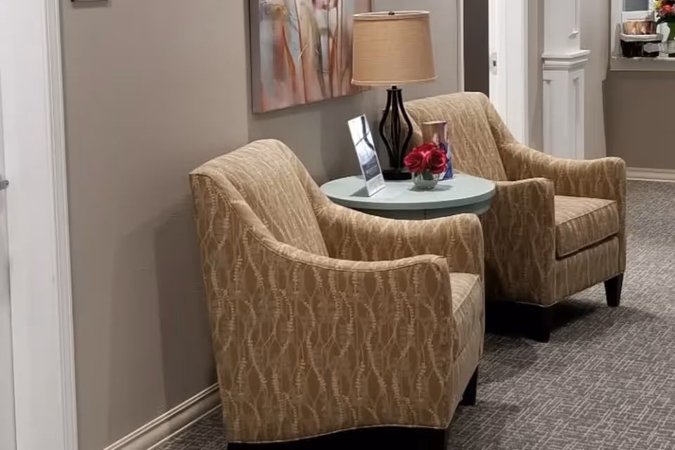 Two upholstered beige armchairs flanking a small round table with a lamp and flowers in a hallway seating area.