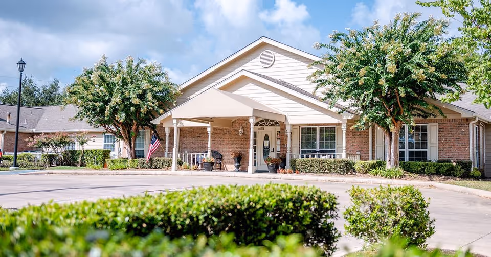 Front exterior view of a single-story senior living facility building with a covered entrance, brick and siding facade, surrounded by trees and shrubs under a partly cloudy sky.