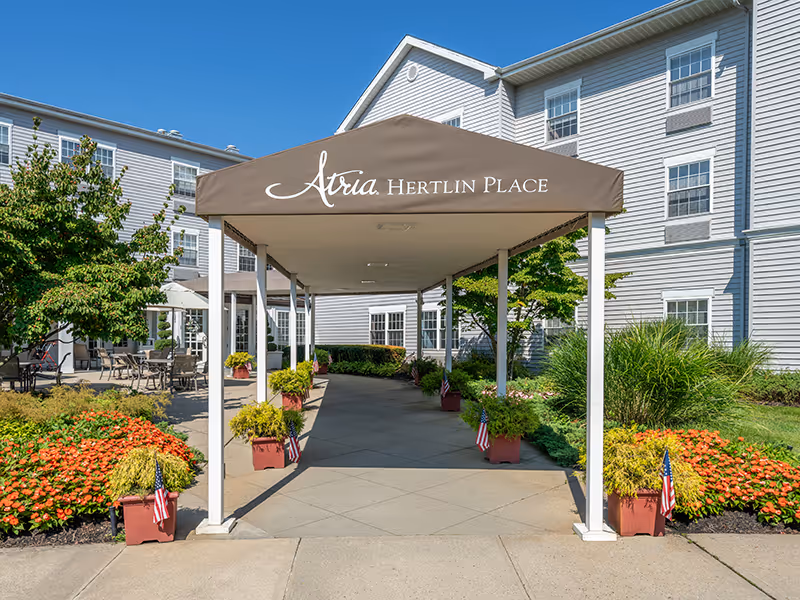 Covered entrance canopy labeled "Atria Hertlin Place" leading to the front of a white multi-story building with landscaped planters.