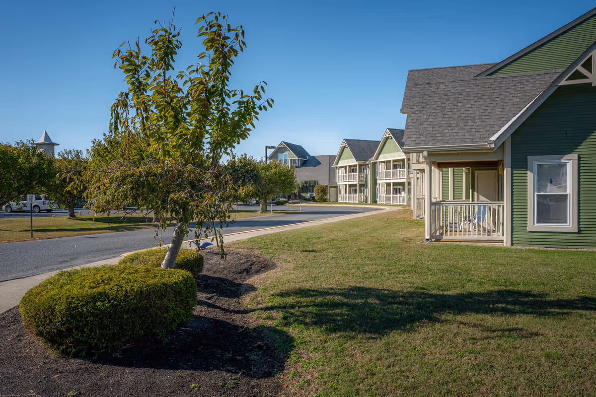 View of a senior living facility exterior with green buildings featuring porches, a paved road, manicured bushes, a small tree, and a clear blue sky.