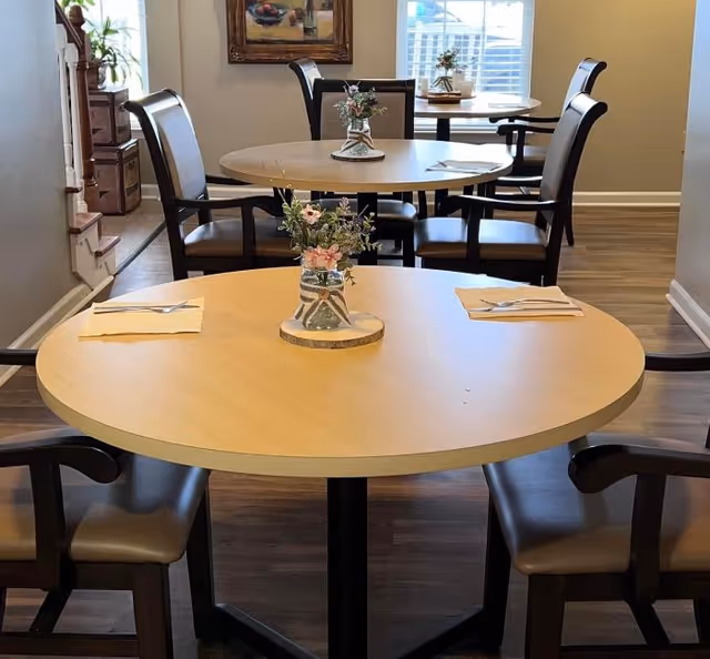A dining area with round wooden tables and black cushioned chairs. Each table has a small floral centerpiece in a glass jar placed on a wooden coaster. Napkins and utensils are neatly set on the tables. The room has wooden flooring and a window with blinds in the background, along with a framed painting on the wall.