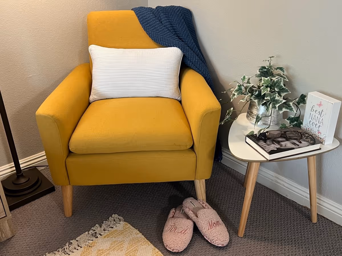 A cozy corner featuring a mustard yellow armchair with a white rectangular pillow and a dark blue knitted throw draped over the back. Next to the chair is a small white side table with wooden legs, holding a potted green plant, a pair of eyeglasses resting on a black and white photo book, and a decorative sign that reads 'best NANA ever'. On the floor in front of the chair are pink slippers with the words 'bird' and 'Mom' embroidered on them. The setting is indoors with a gray carpet and a beige wall.