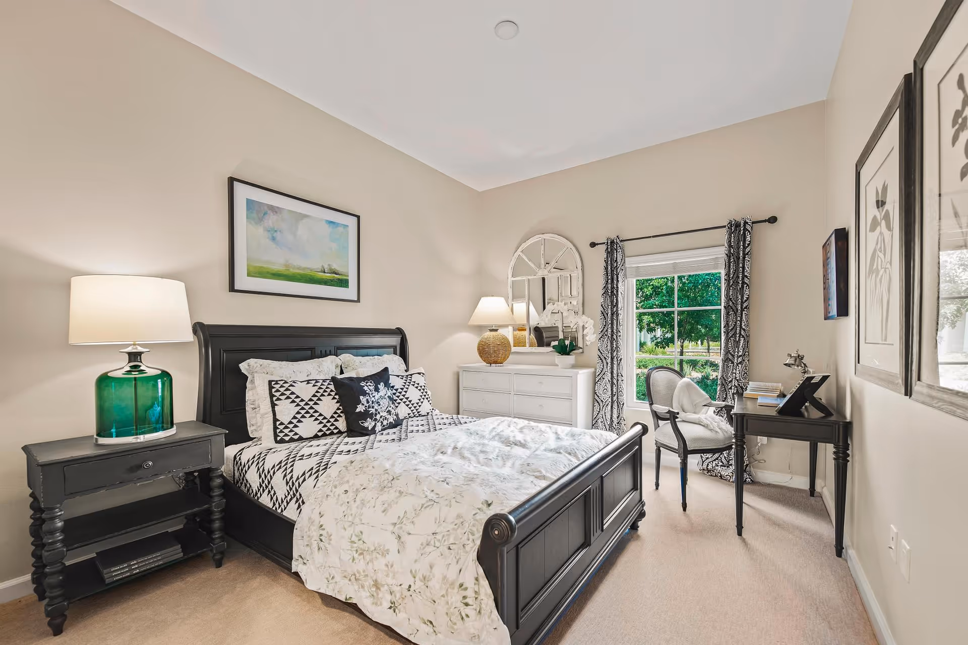 A cozy bedroom with a black wooden bed frame, patterned black and white bedding, and a green glass lamp on a black nightstand. There is a white dresser with a round mirror and a lamp on top, a window with patterned curtains, and a small black desk with a chair. The walls are beige and decorated with framed artwork.