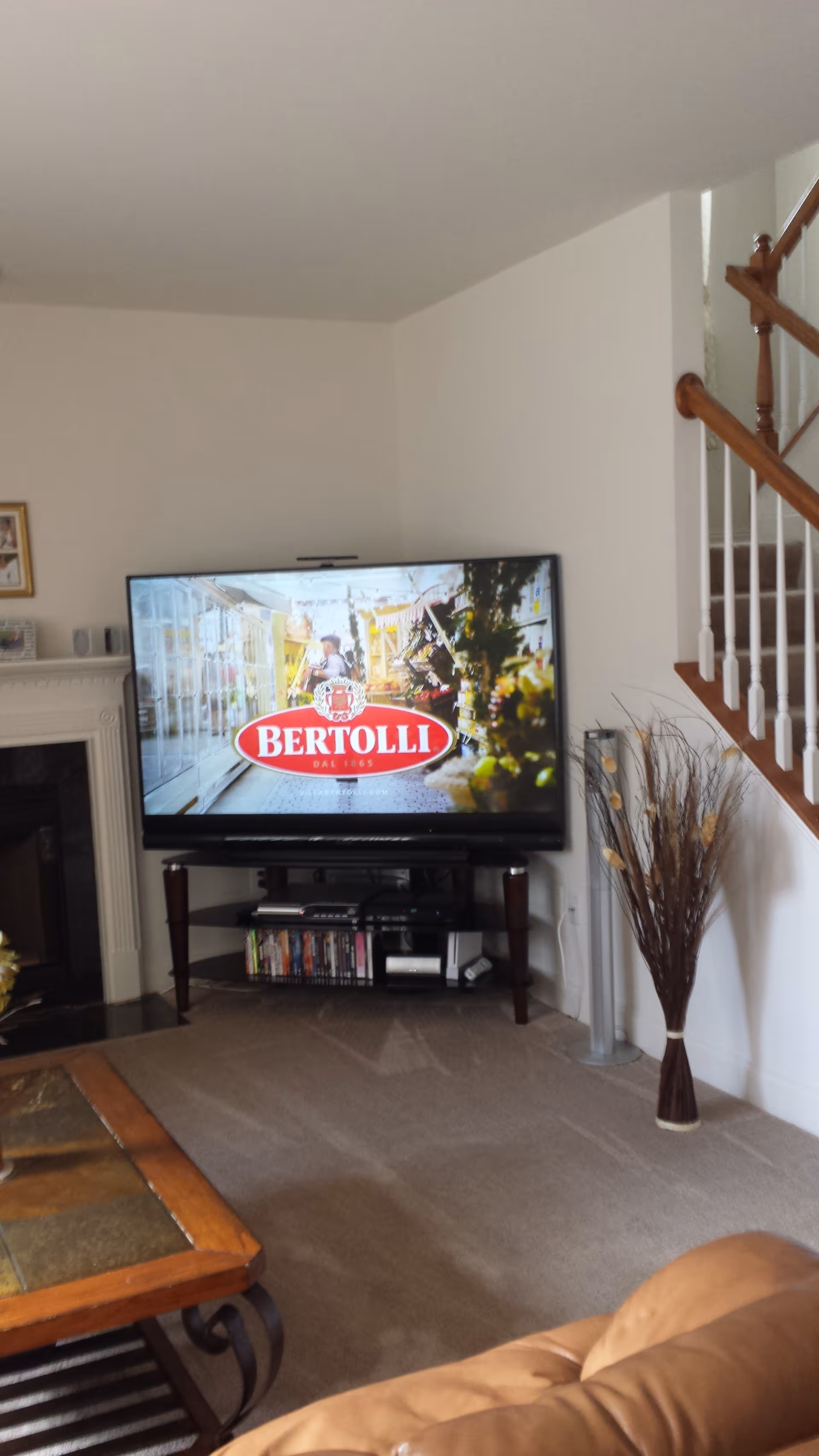 A cozy living room with a flat-screen TV displaying a Bertolli advertisement. The TV is on a dark wooden stand with shelves holding DVDs and electronic devices. To the right is a staircase with wooden handrails and white balusters. A tall vase with decorative branches stands near the staircase. A coffee table with a tiled top and a brown leather couch are partially visible in the foreground. The walls are painted white, and there is a fireplace on the left side of the room.