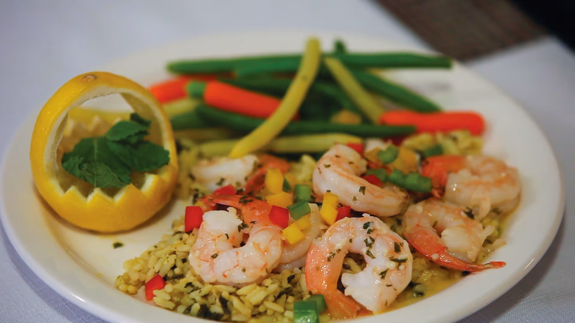 A plate of cooked shrimp served on a bed of seasoned rice with diced red, yellow, and green bell peppers, accompanied by steamed green, yellow, and red beans, and garnished with a lemon half carved into a basket shape holding fresh mint leaves.