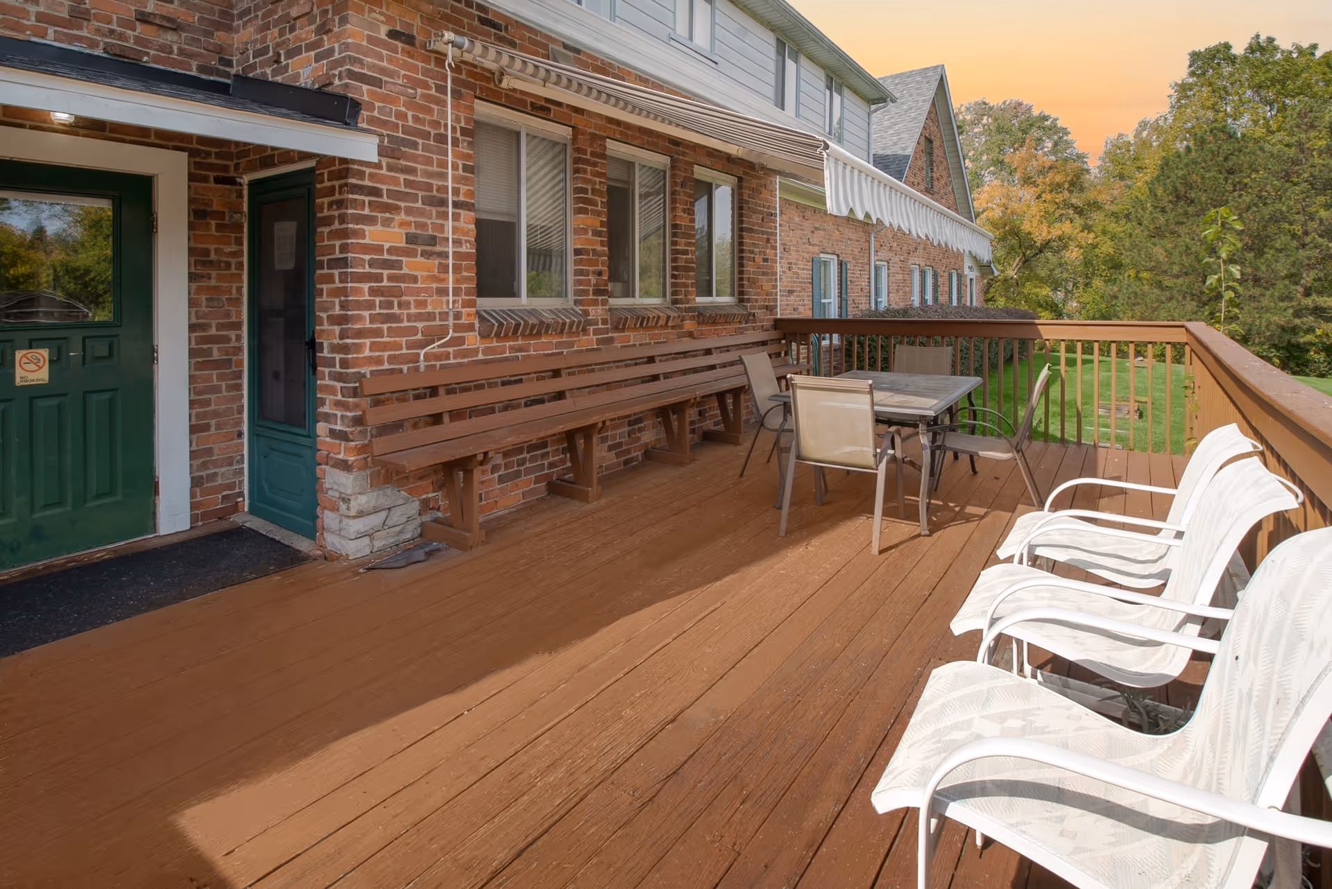 Outdoor wooden deck attached to a brick building with green doors and windows. The deck has a long wooden bench along the wall, a table with four chairs, and three white chairs lined up along the railing. Trees and greenery are visible in the background under a sunset sky.