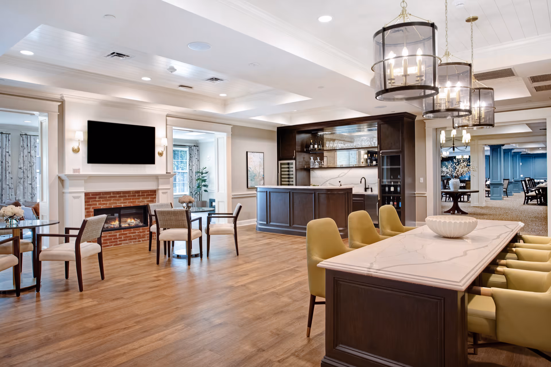 A spacious and well-lit common area with wooden flooring, featuring a modern fireplace with a TV mounted above it, several small tables with chairs, a bar area with glass shelves and wine bottles, and a large marble-topped table with yellow upholstered chairs. The ceiling has recessed lighting and three large hanging lantern-style light fixtures.